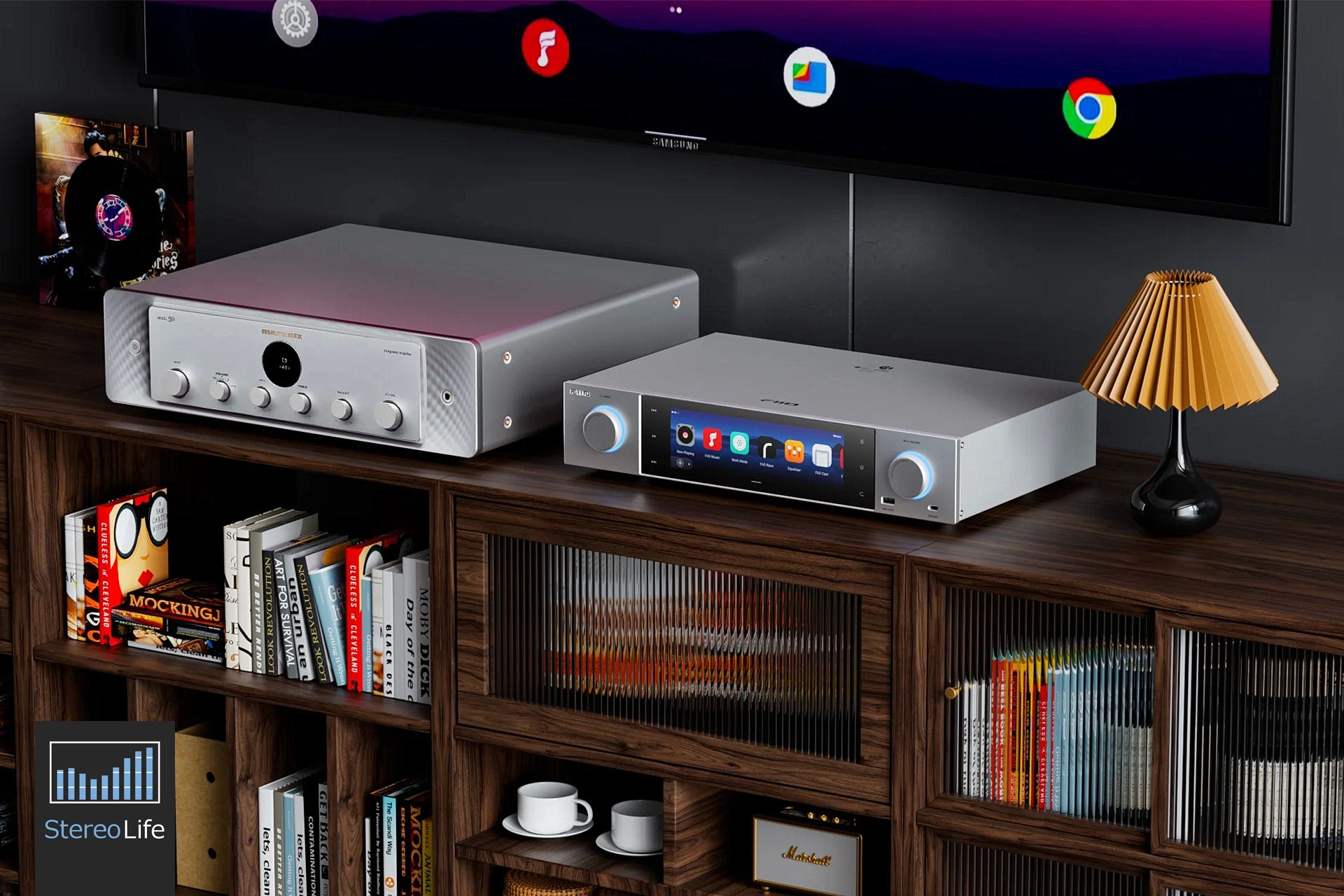 Dos dispositivos de audio plateados con pantallas sobre un mueble de madera lleno de libros, con un televisor arriba.