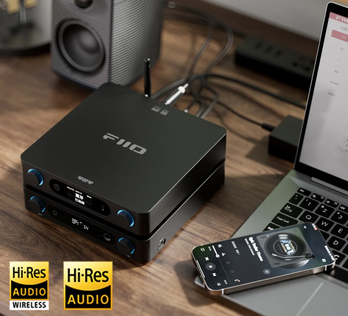 Czarne urządzenie audio FiiO na drewnianym biurku z głośnikiem, laptopem i smartfonem.