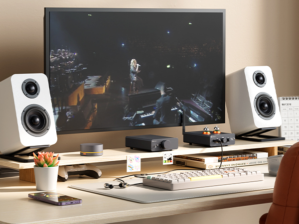 Ein Schreibtisch mit Monitor, weißen Lautsprechern, Audiogeräten, einer Tastatur und einem kleinen Pflanzentopf.