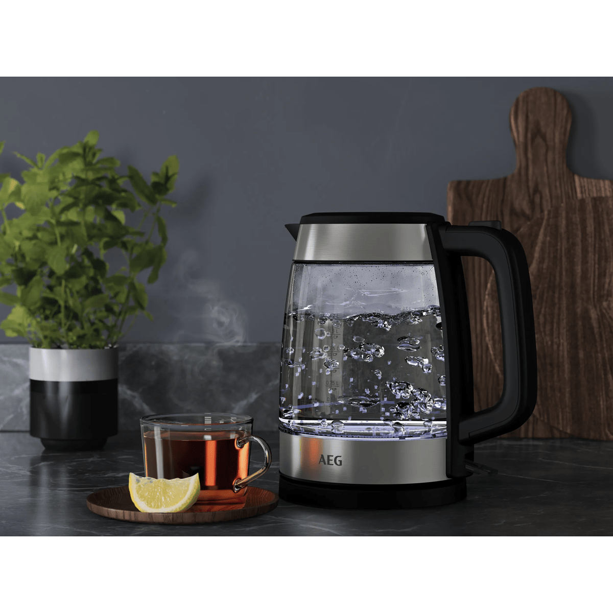 Un bollitore in vetro AEG illuminato bolle acqua vicino a una tazza di tè, limone, pianta in vaso e tagliere.