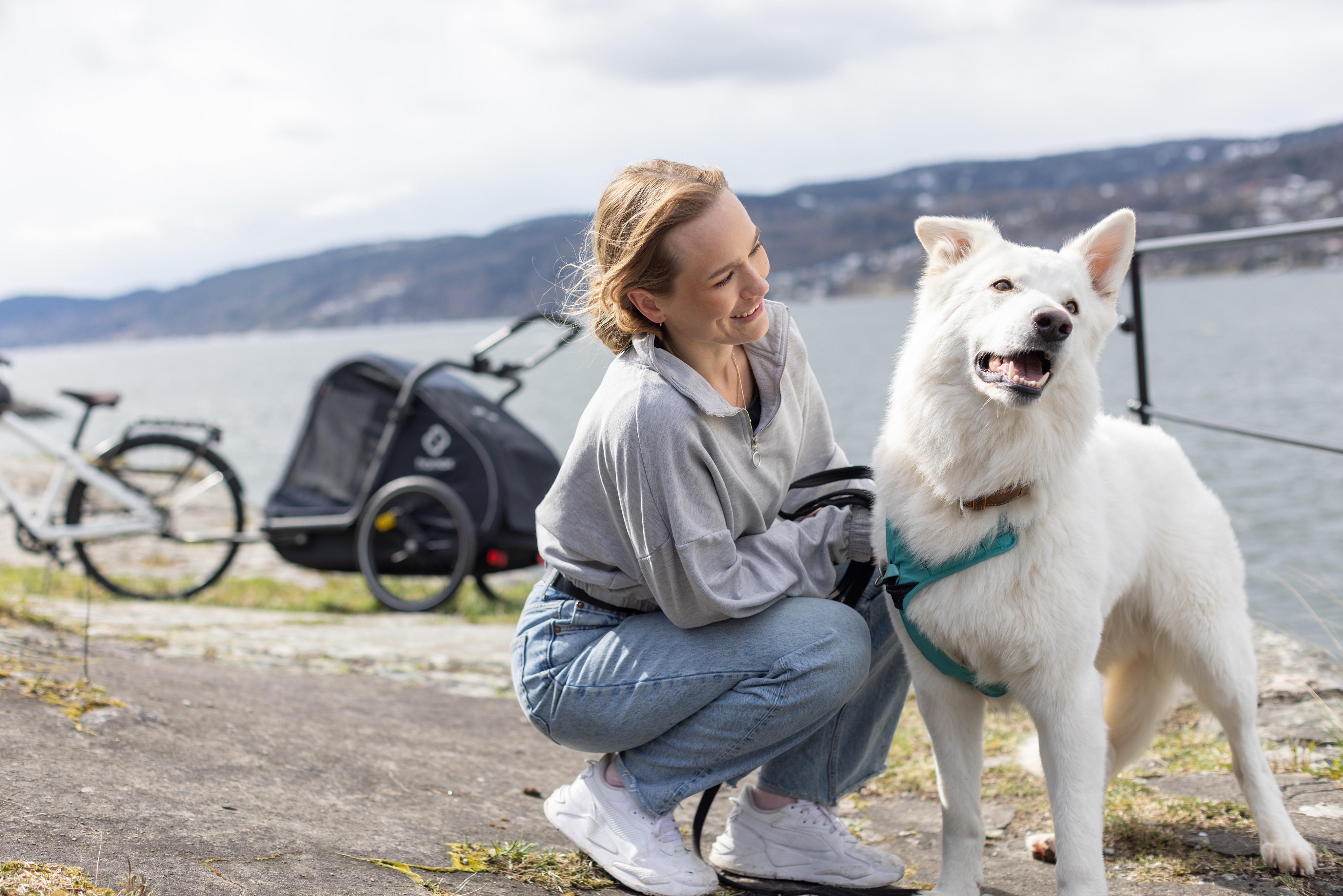Una mujer sonriente se agacha junto a un perro blanco cerca del agua, con bicicleta y remolque al fondo.
