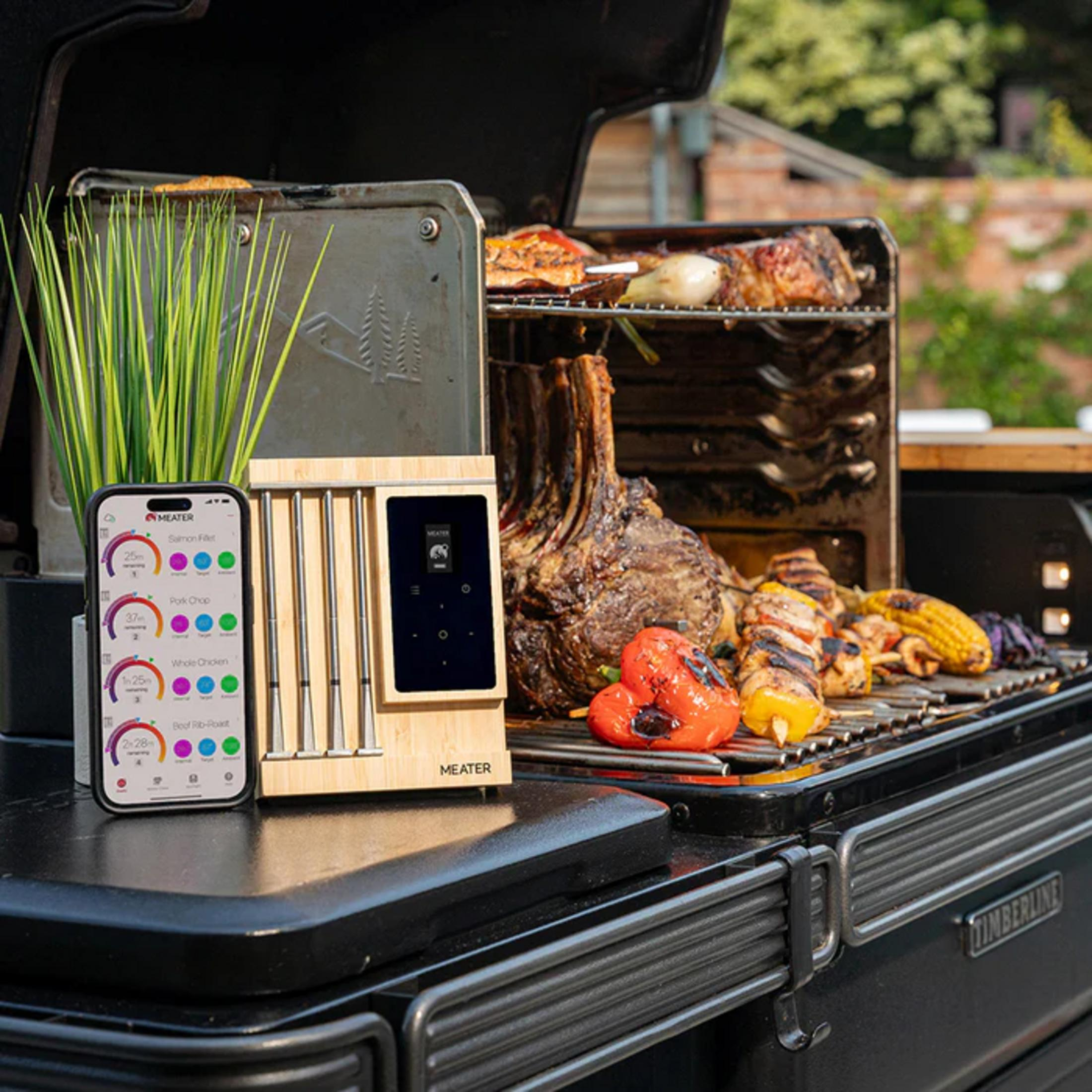 Un smartphone et un thermomètre à viande sans fil devant un gril fumant rempli de viande et de légumes.