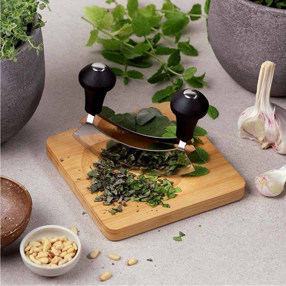 Ein Wiegemesser hackt Kräuter auf einem Holzbrett, umgeben von Pinienkernen, Knoblauch und Blumentöpfen.