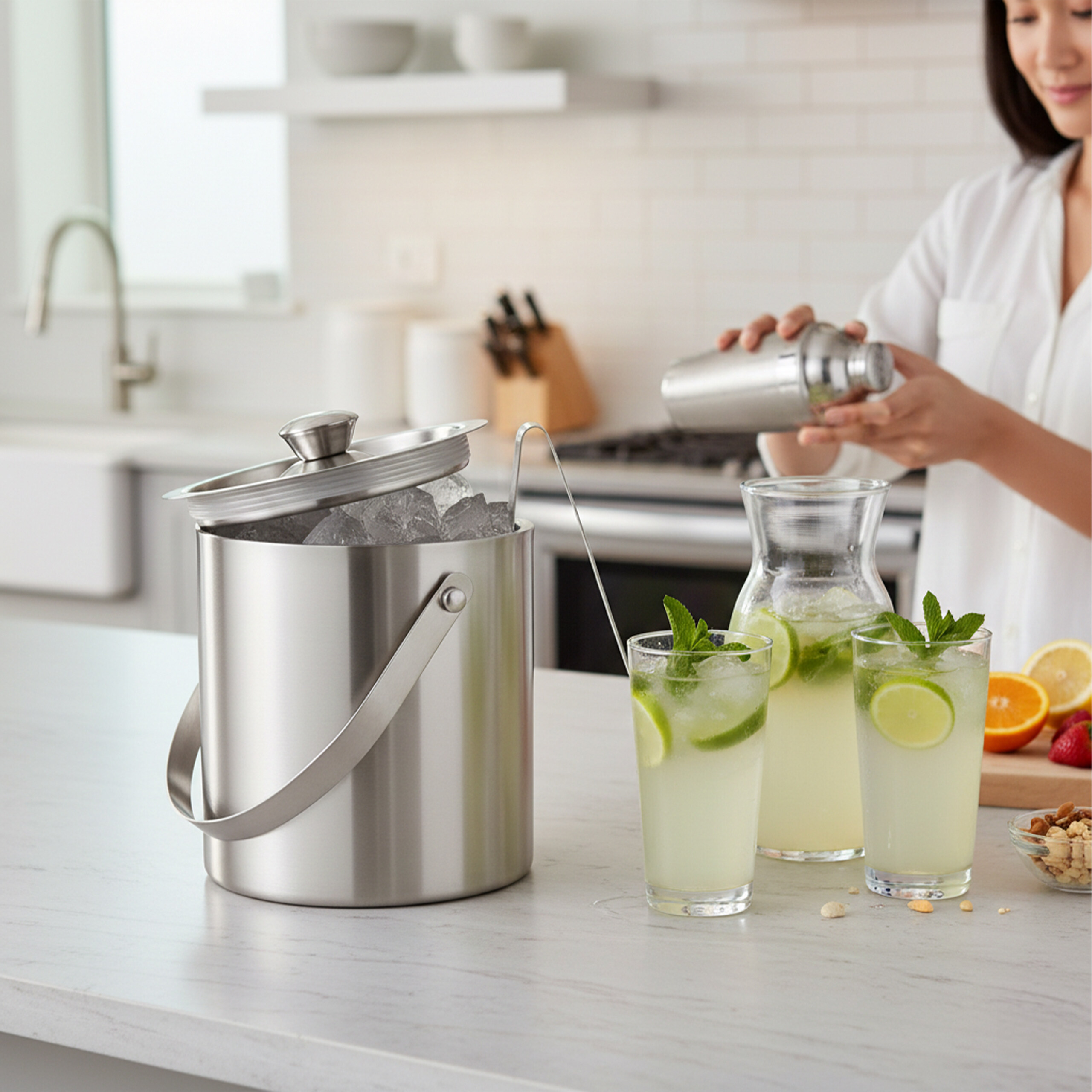 Edelstahl-Eiseimer und Gläser mit Limonade auf einer Küchentheke, während eine Frau im Hintergrund einen Shaker hält.