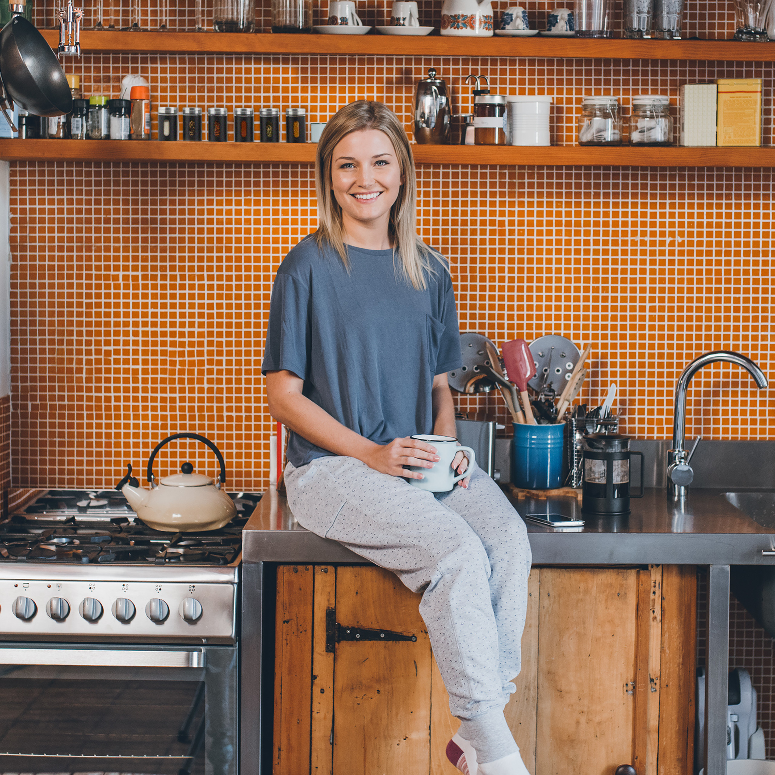 Eine blonde Frau in grauem Shirt und gemusterten Hosen sitzt auf einer Küchentheke, hält eine Tasse und lächelt.