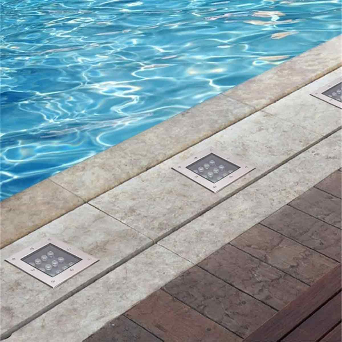 Quadratische Einbaustrahler in Steinfliesen am Rand eines klaren blauen Pools.