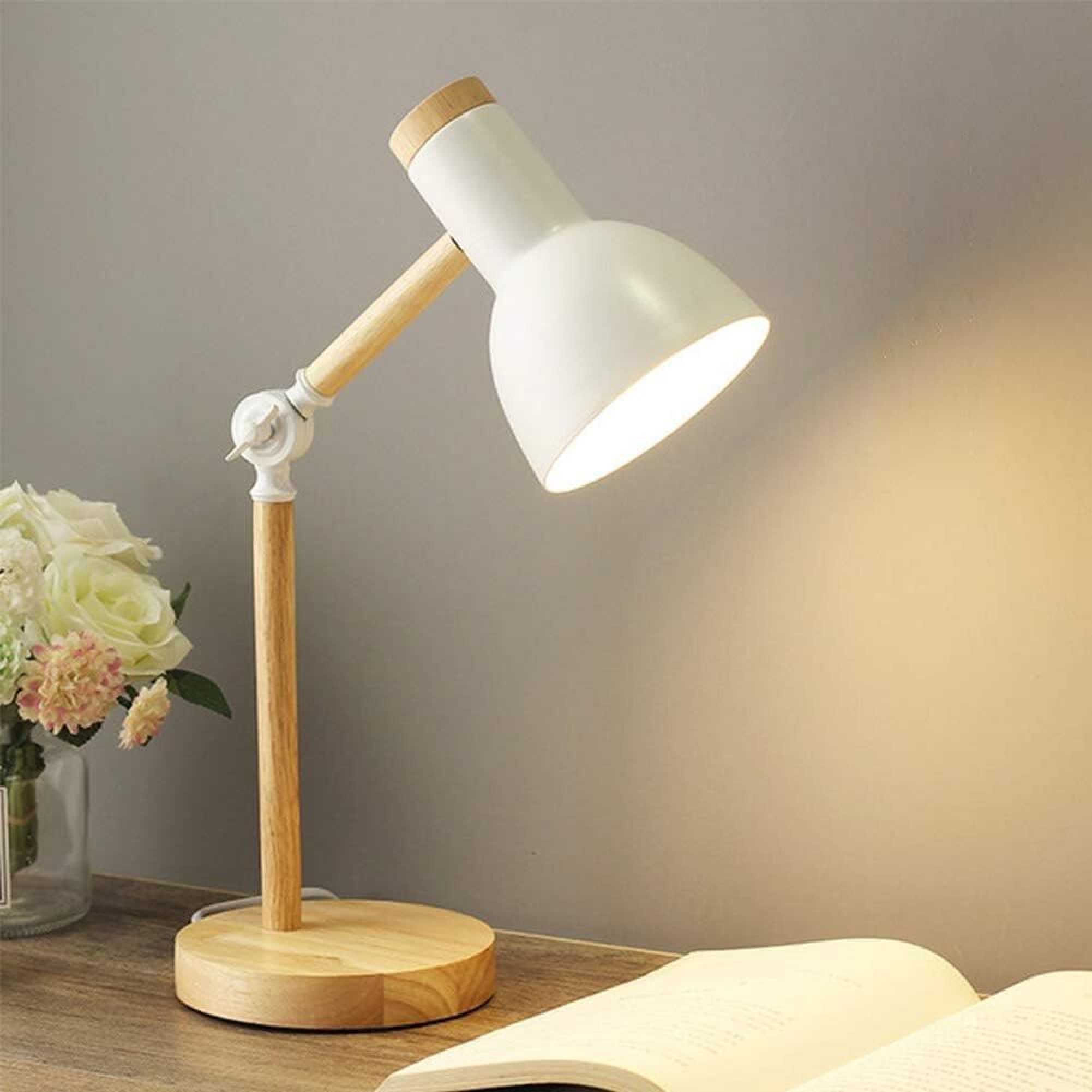 Eine weiße und hölzerne Schreibtischlampe brennt neben einem offenen Buch und einer Blumenvase.