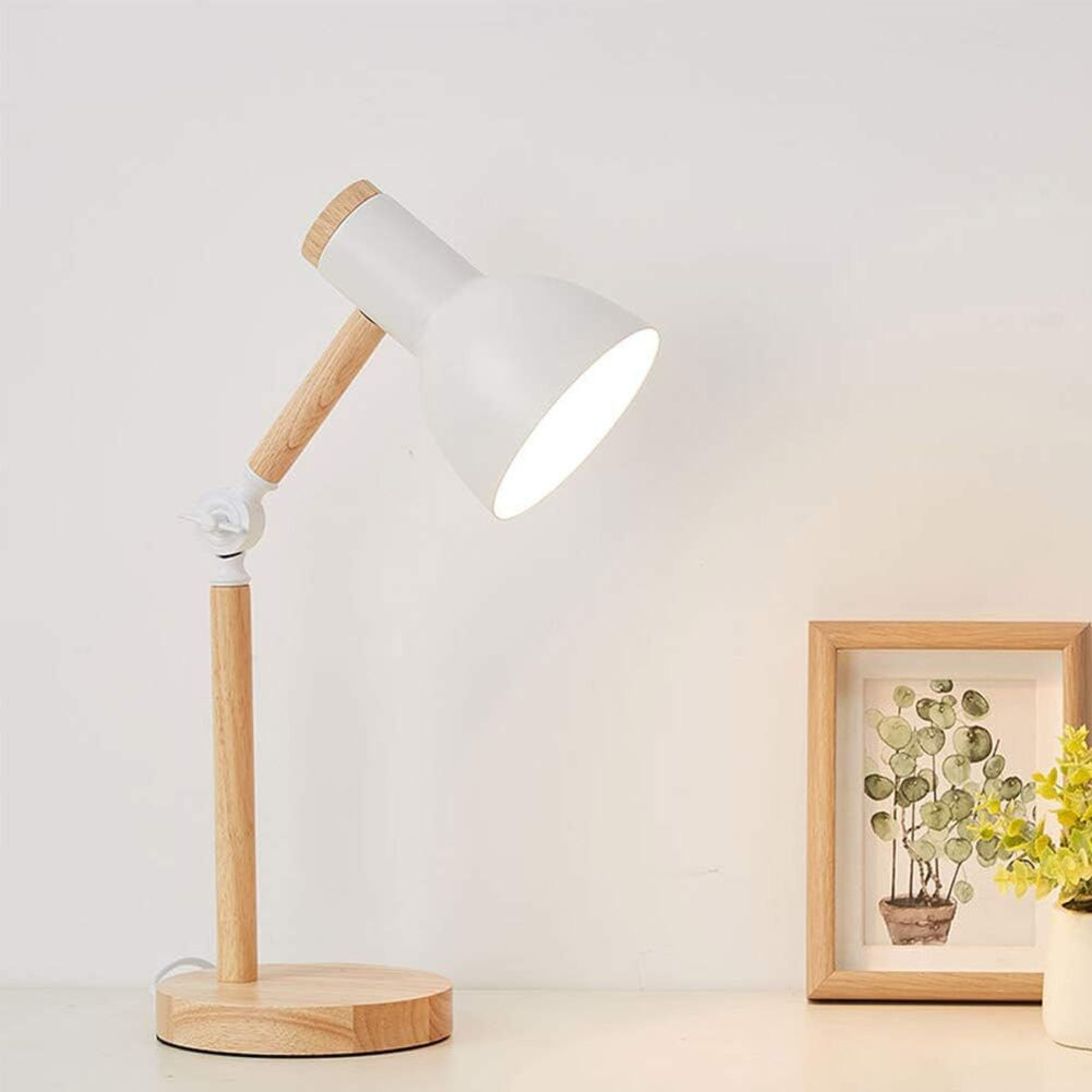 Eine weiße und hölzerne Schreibtischlampe leuchtet auf einem Tisch, daneben ein gerahmtes Pflanzenbild.