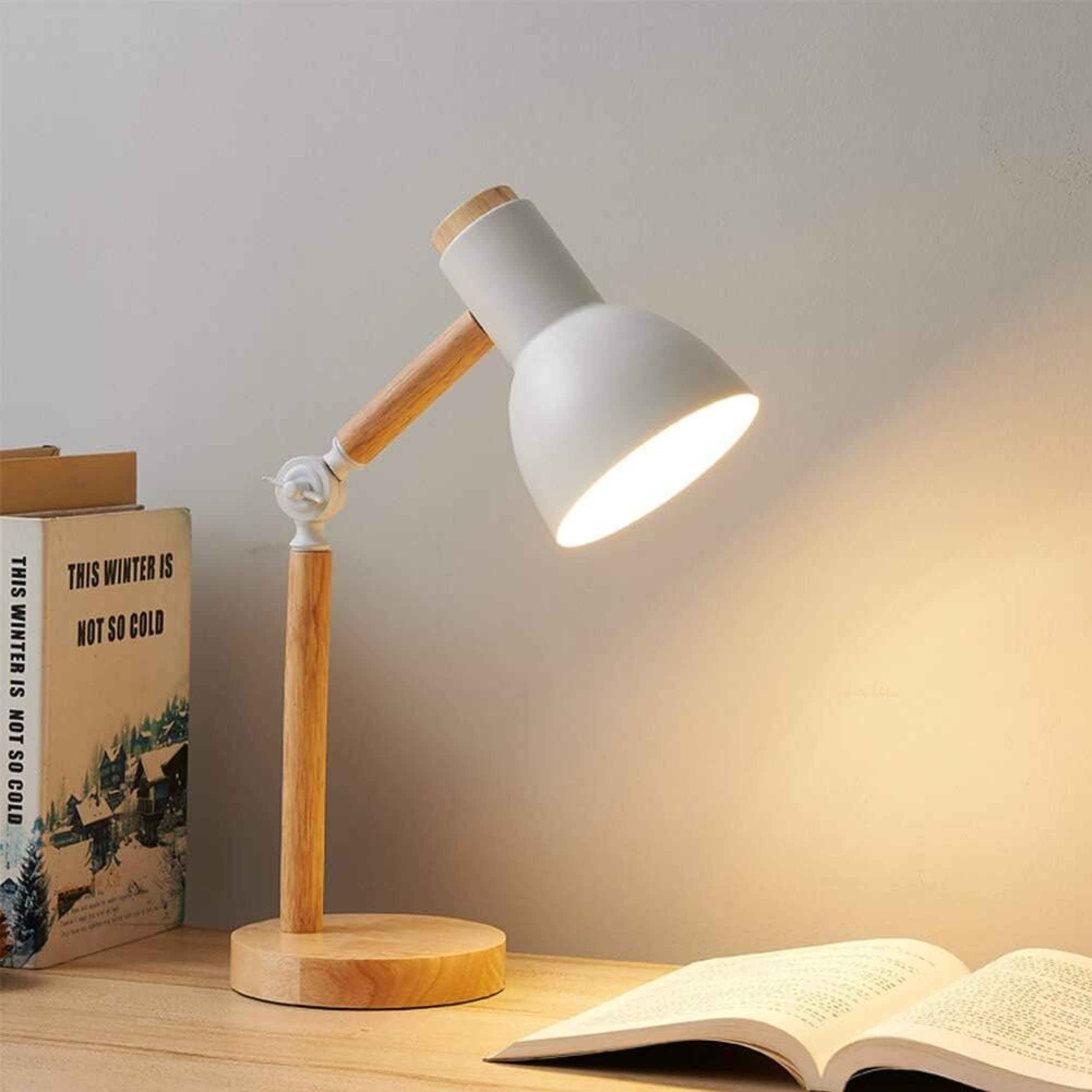 Eine weiße und hölzerne Schreibtischlampe leuchtet neben offenen Büchern auf einem Holztisch vor einer grauen Wand.