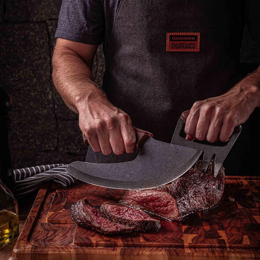 Eine Person in einer dunklen Schürze schneidet gekochtes Steak mit einem Wiegemesser auf einem Holzbrett.