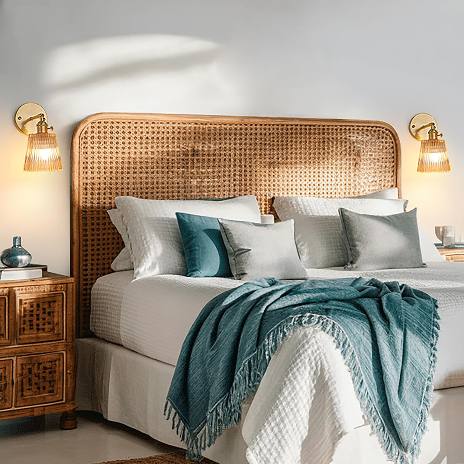 Gemütliches Schlafzimmer mit Rattan-Kopfteil, weißem Bett, blauer Decke, Kissen und Wandleuchten.