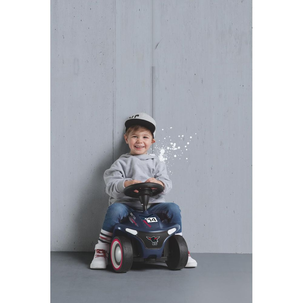 Ein kleiner Junge lächelt, während er auf einem blauen Rutschauto vor einer grauen Wand sitzt.