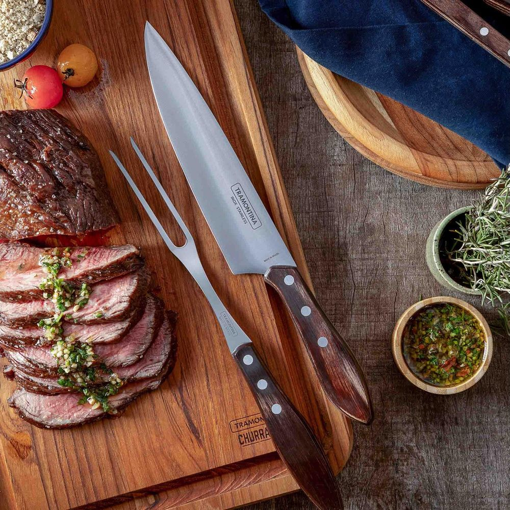 Ein großes Fleischmesser und eine Fleischgabel auf einem Holzbrett neben tranchiertem Fleisch und Kräutern.