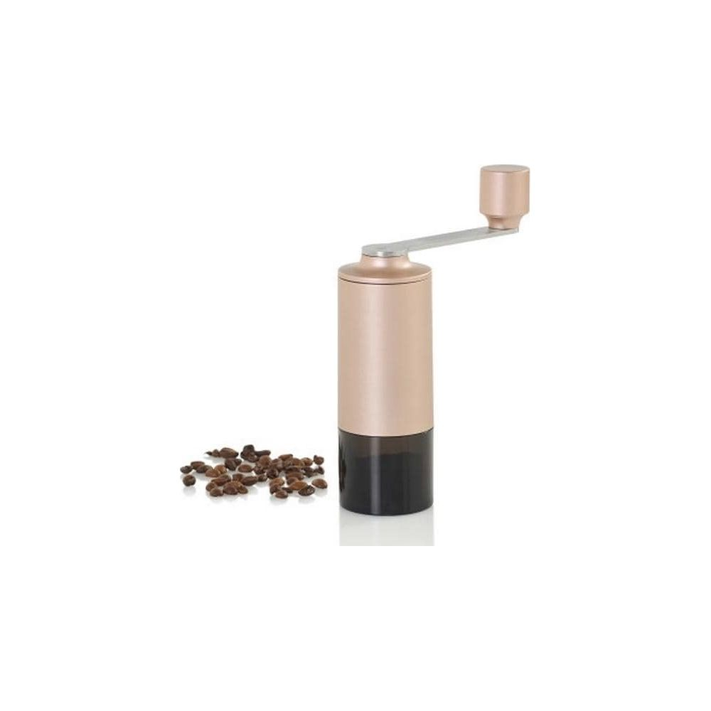 Un macinacaffè manuale in oro rosa si trova su sfondo bianco accanto a un piccolo mucchio di chicchi di caffè.