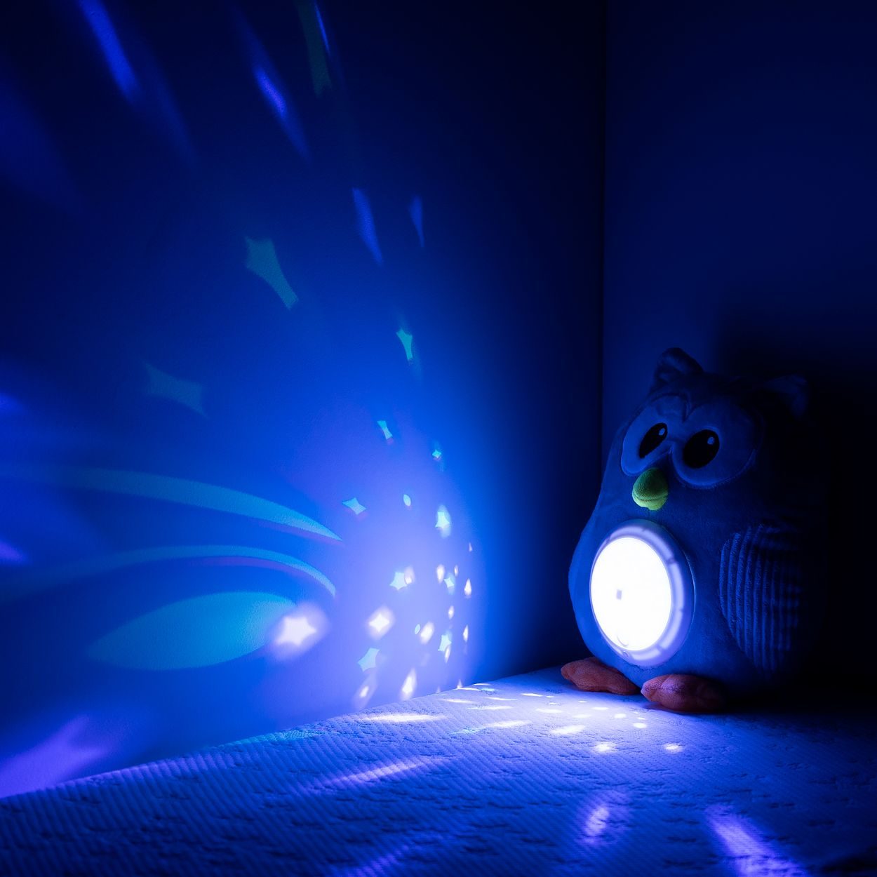 Ein blaues Eulen-Nachtlicht sitzt auf einem Bett und projiziert Sternen- und Lichtmuster an die dunkle Wand.
