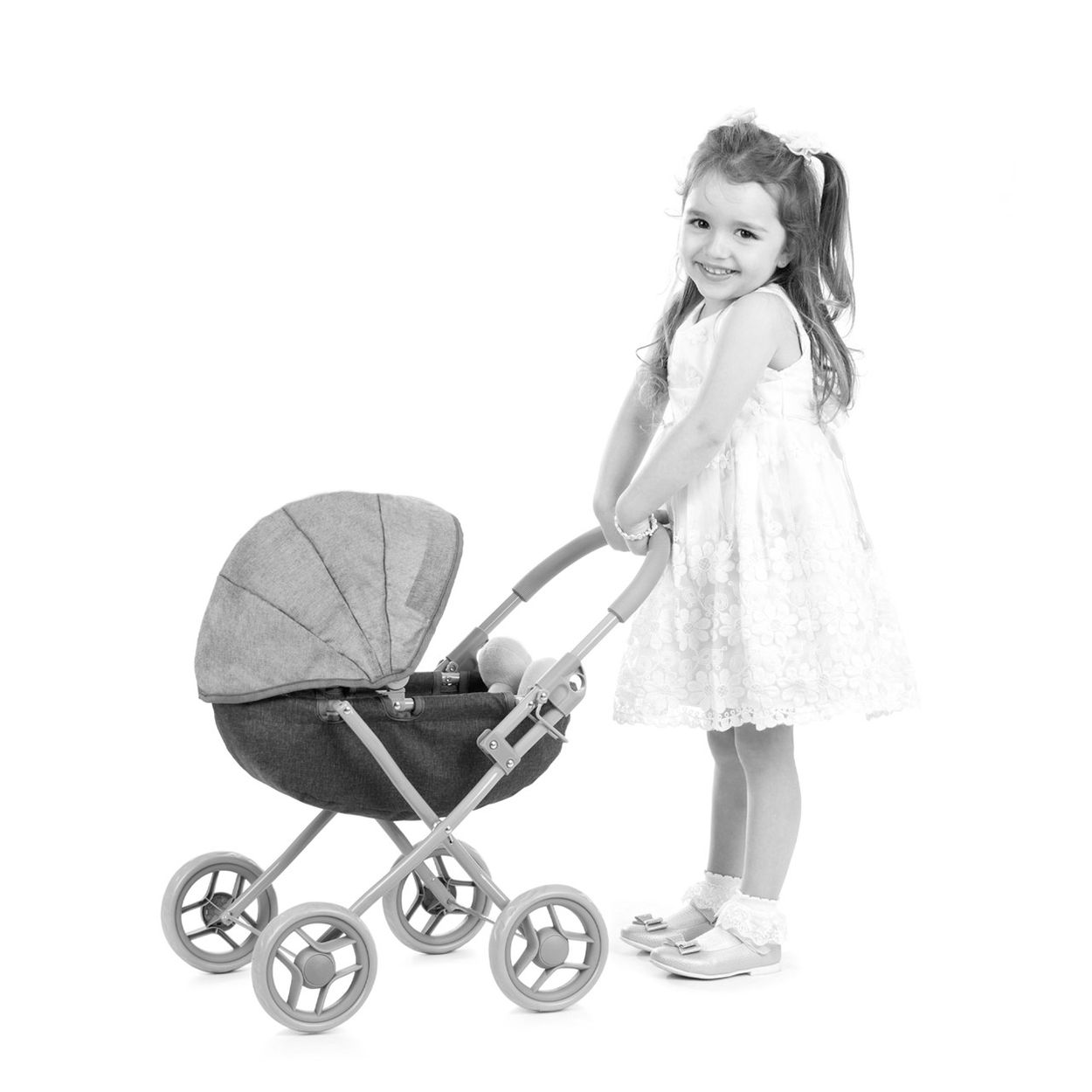 Ein kleines Mädchen in einem Sommerkleid lächelt in die Kamera und schiebt einen kleinen Spielzeug-Kinderwagen.