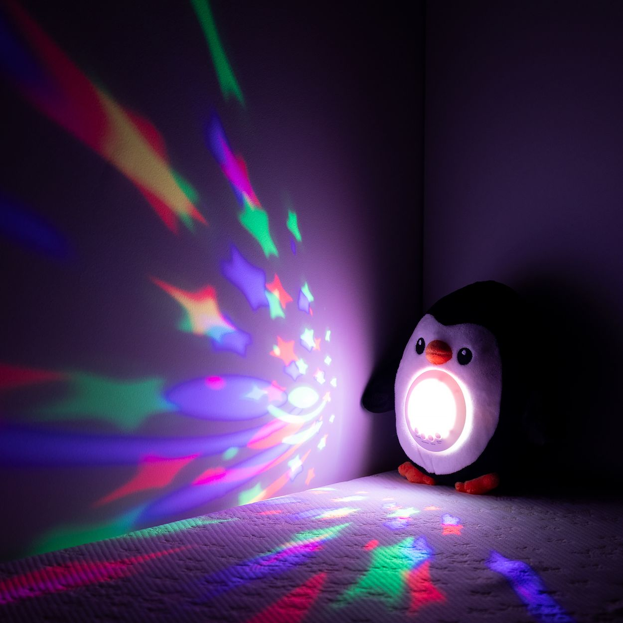 Ein Plüschpinguin mit hellem Bauch projiziert bunte Sterne und Lichtstrahlen an die Wand.