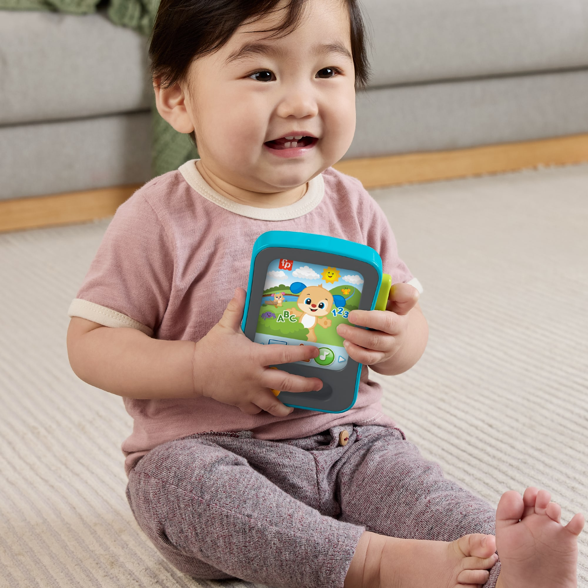 Ein lächelndes Baby sitzt auf einem Teppich und hält ein buntes Spielzeug-Tablet mit einem Welpen.