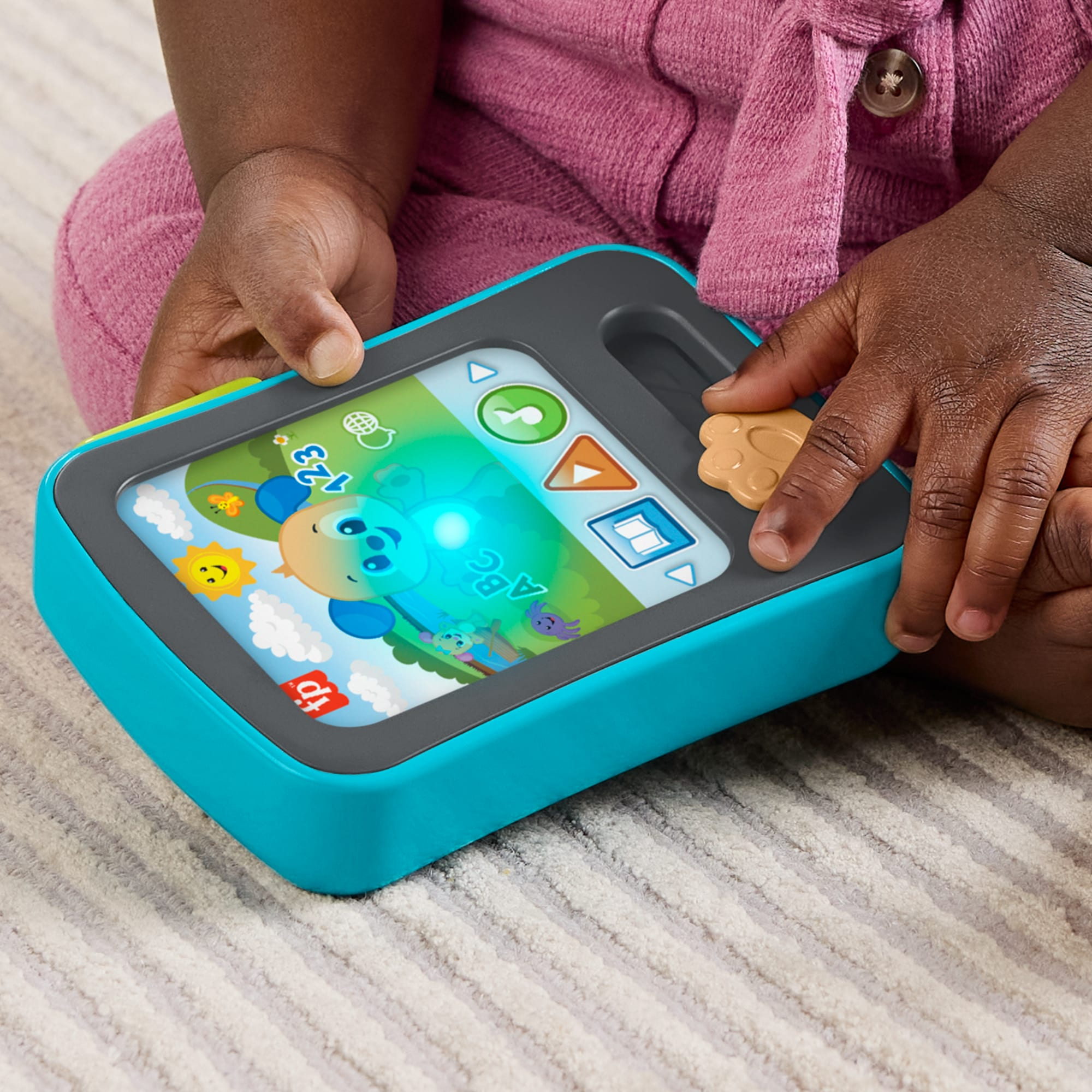 Ein Kleinkind hält ein hellblaues und graues Spielzeugtablet mit leuchtendem Bildschirm auf einem hellen Teppich.