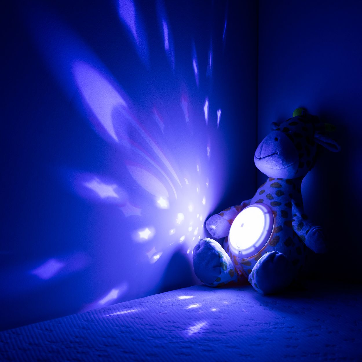 Ein Plüsch-Giraffen-Nachtlicht sitzt in einer Ecke und wirft blaue, sternförmige Lichter an die dunkle Wand.