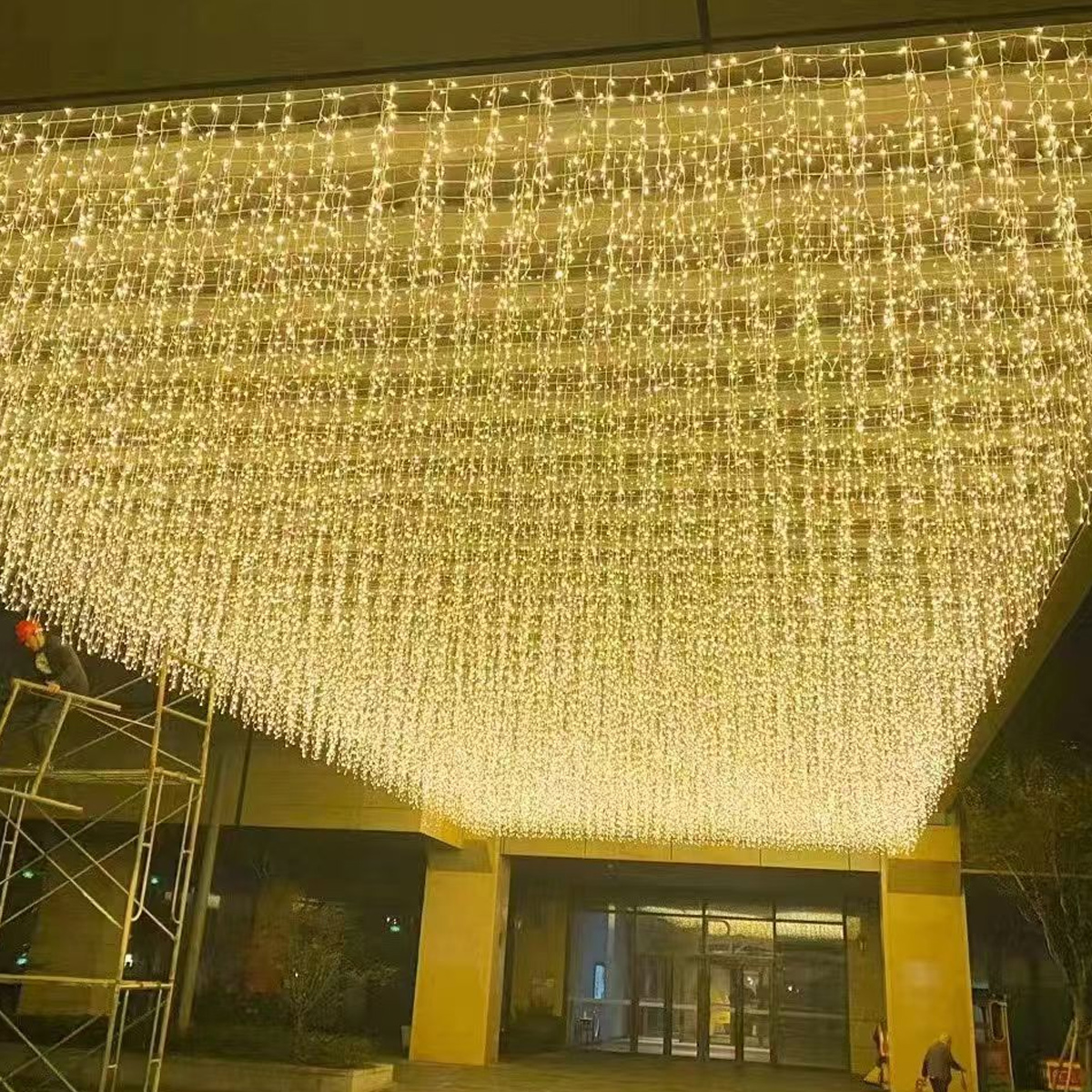 Ein großes Deckenbaldachin aus warmweißen Lichterketten über einem modernen Hoteleingang bei Nacht.