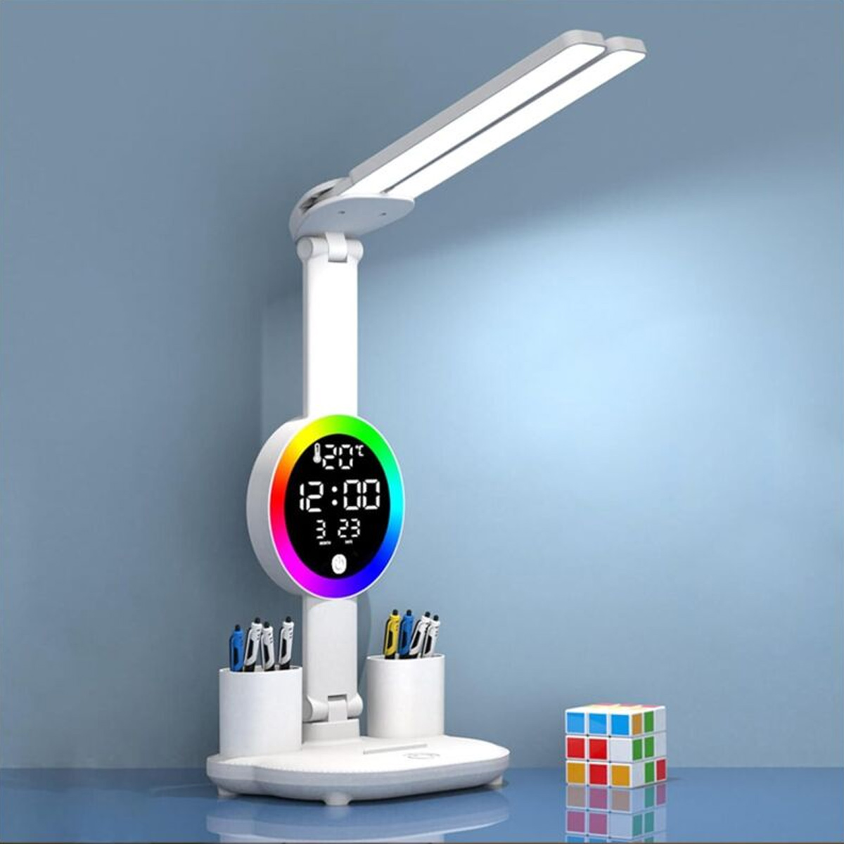 Weiße Schreibtischlampe mit zwei Köpfen, Digitaluhr, Stiftehaltern und einem Zauberwürfel daneben auf blauer Fläche.