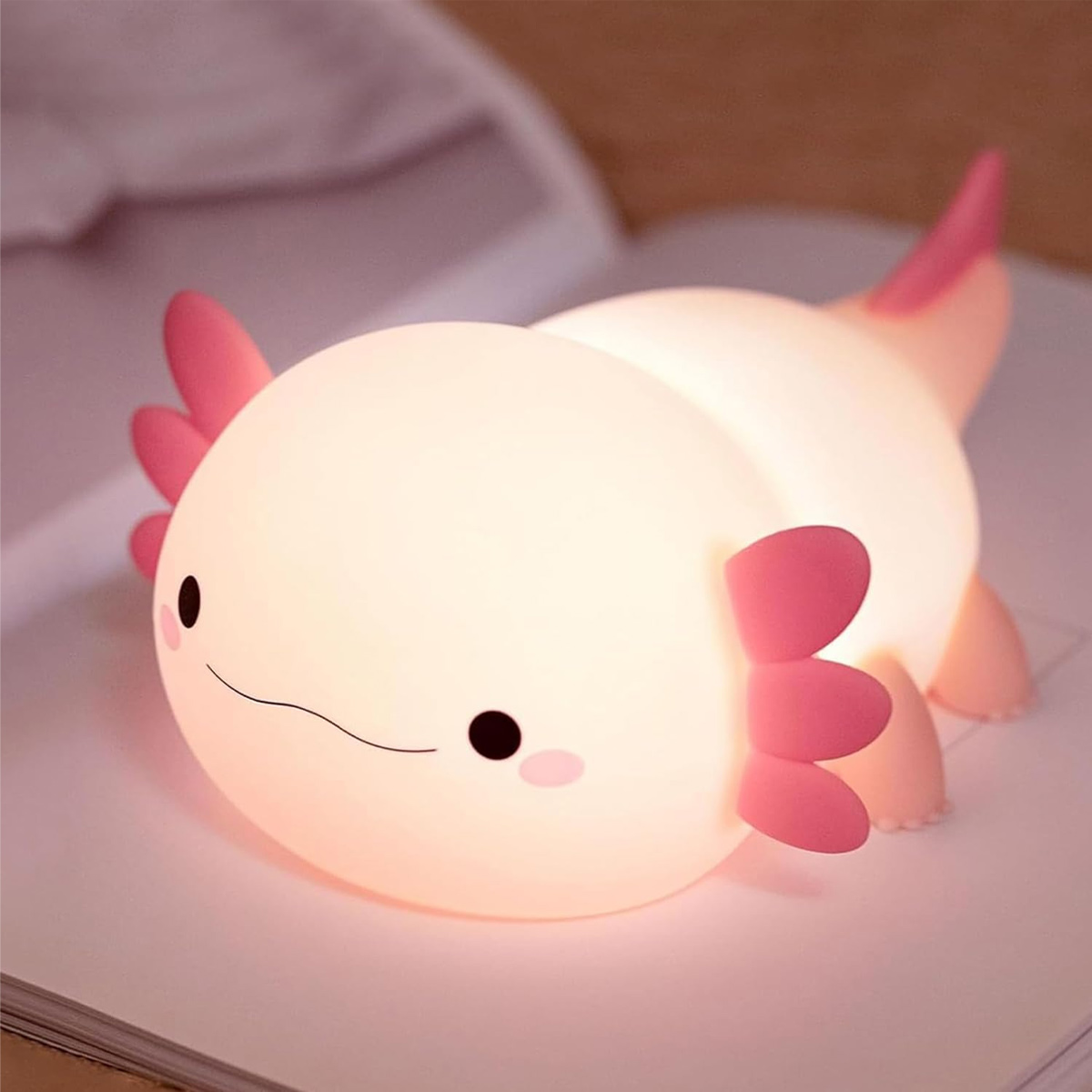 Eine niedliche, hell leuchtende Axolotl-Lampe mit pinken Kiemen sitzt auf einem offenen Buch.