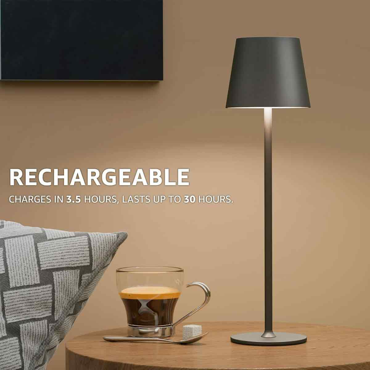 Eine elegante, dunkelgraue Akku-Tischlampe steht auf einem Holztisch neben einer Kaffeetasse und einem Kissen.