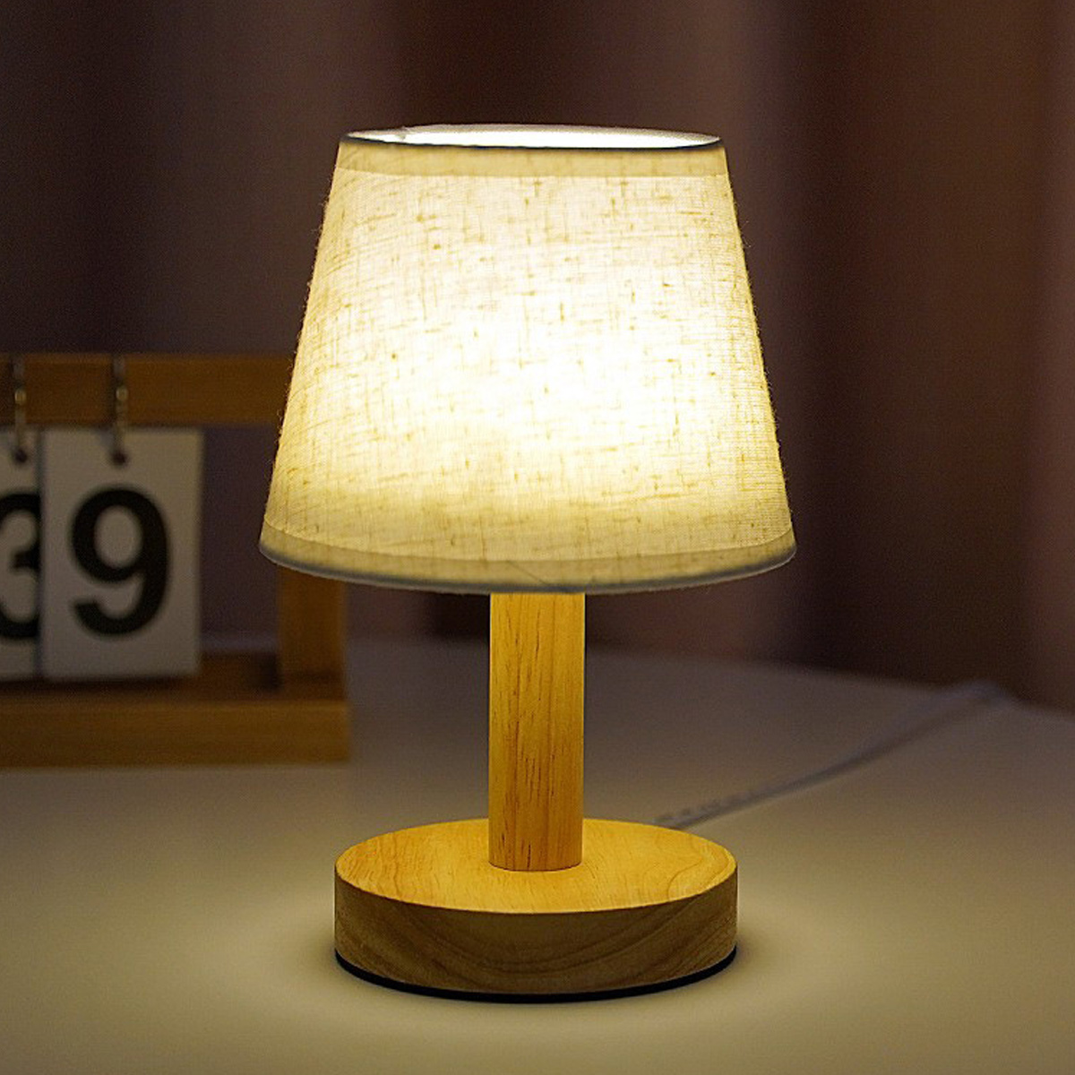 Eine kleine Holztischlampe mit beigem Lampenschirm steht auf einem Tisch in einem warmen Raum.