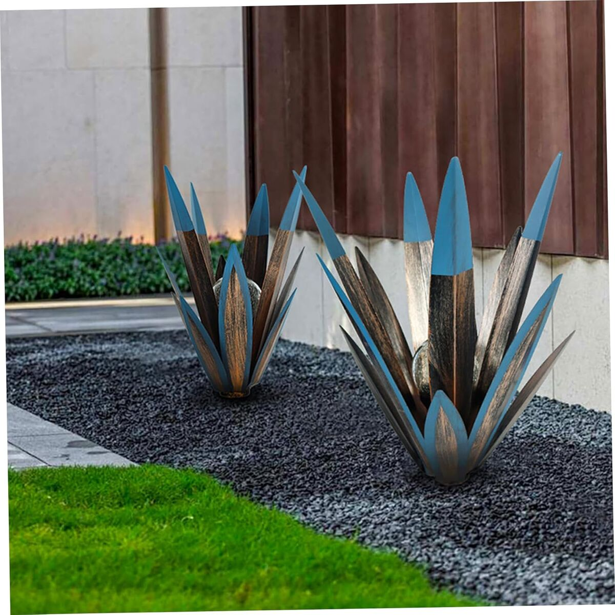 Zwei Agavenskulpturen aus Metall mit blauen Spitzen stehen in einem Kiesbeet vor einem Gebäude.