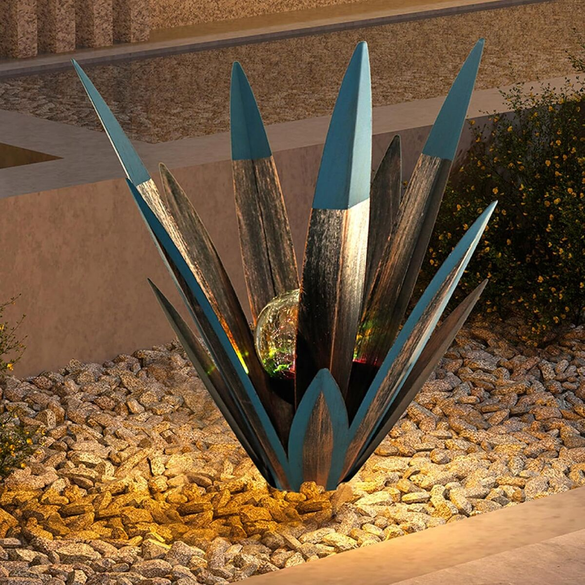 Eine agave-ähnliche Metallskulptur mit blau-kupfernen Blättern und einer gläsernen Kugel im Zentrum, auf Kieselsteinen.