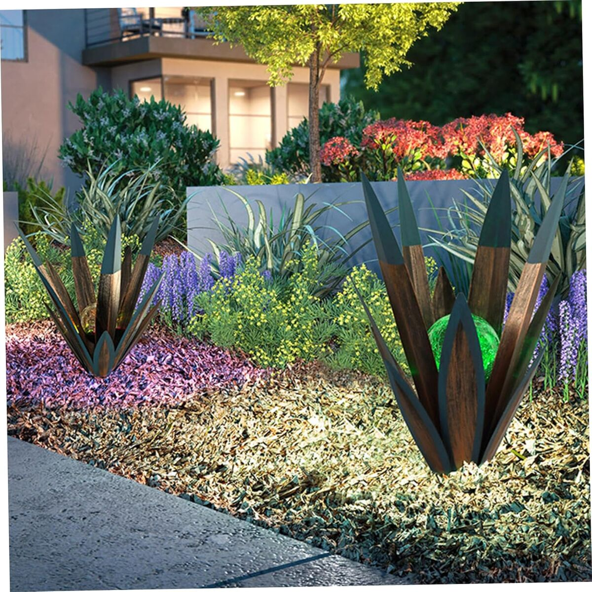 Zwei agaveförmige Metallskulpturen mit leuchtenden grünen Kugeln in einem farbenfrohen Garten.