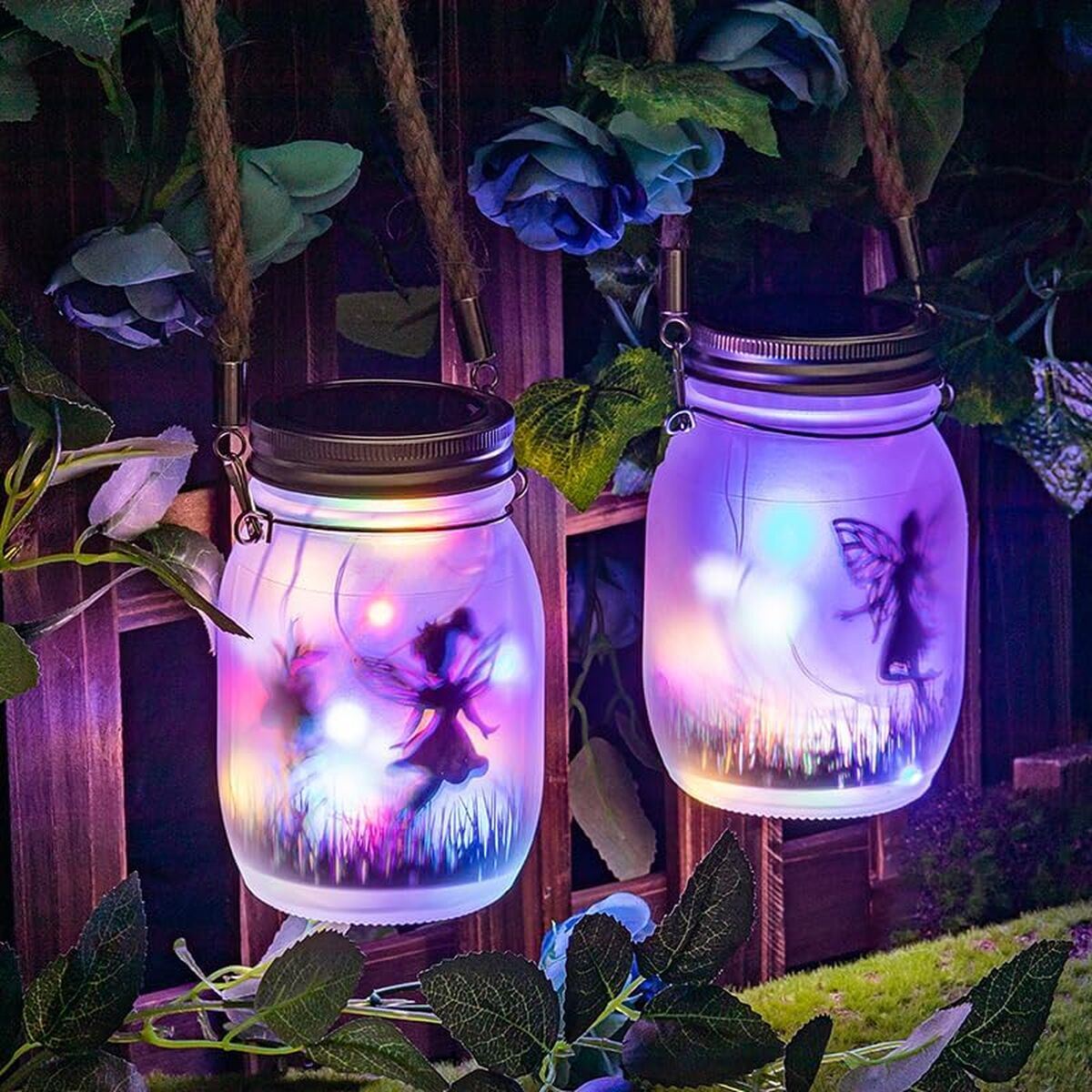 Zwei hängende Einmachgläser mit Lichterketten und Feenmotiven, umgeben von künstlichen blauen Rosen und grünem Laub.