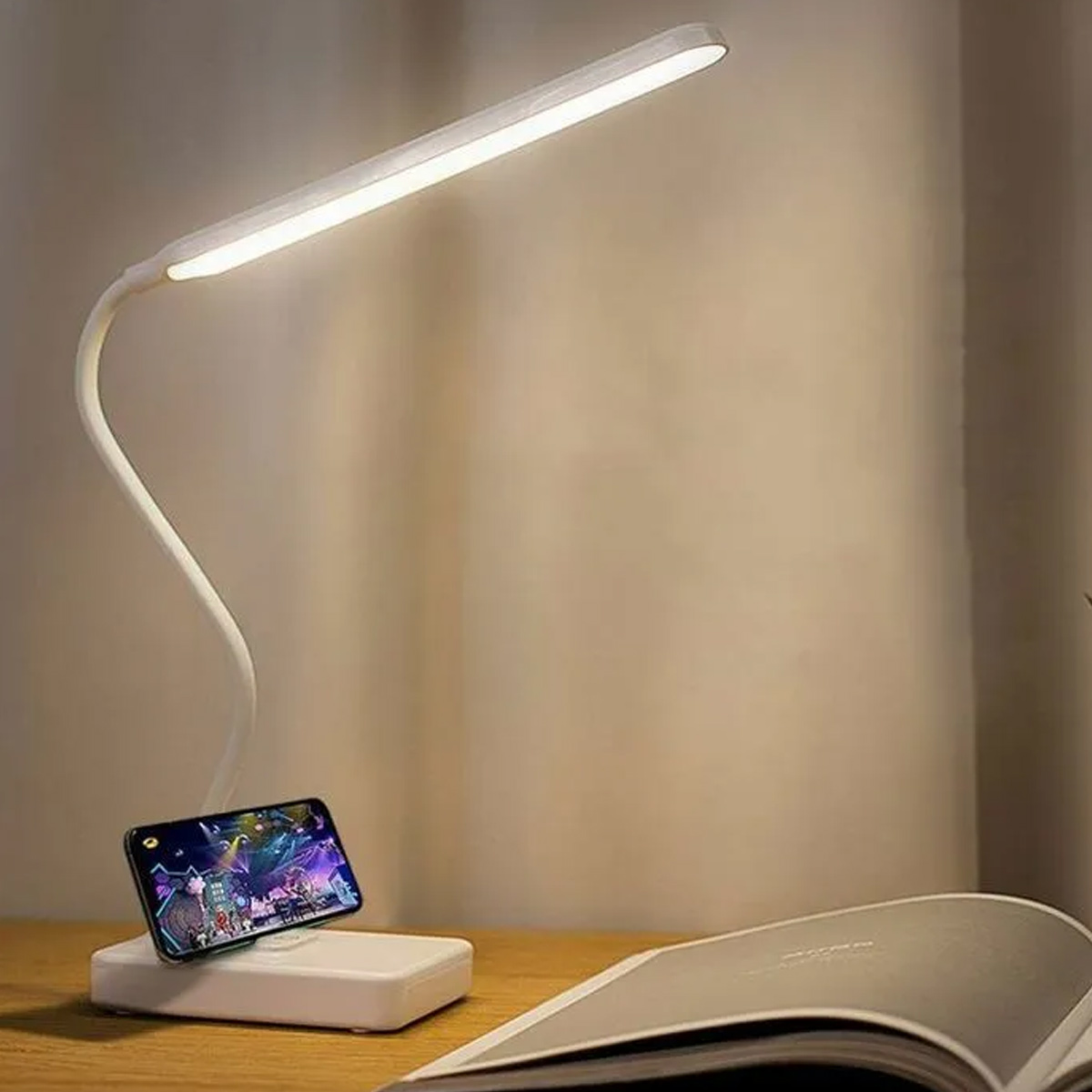 Eine weiße LED-Schreibtischlampe mit flexiblem Hals und Handyständer neben einem offenen Buch auf einem Holztisch.