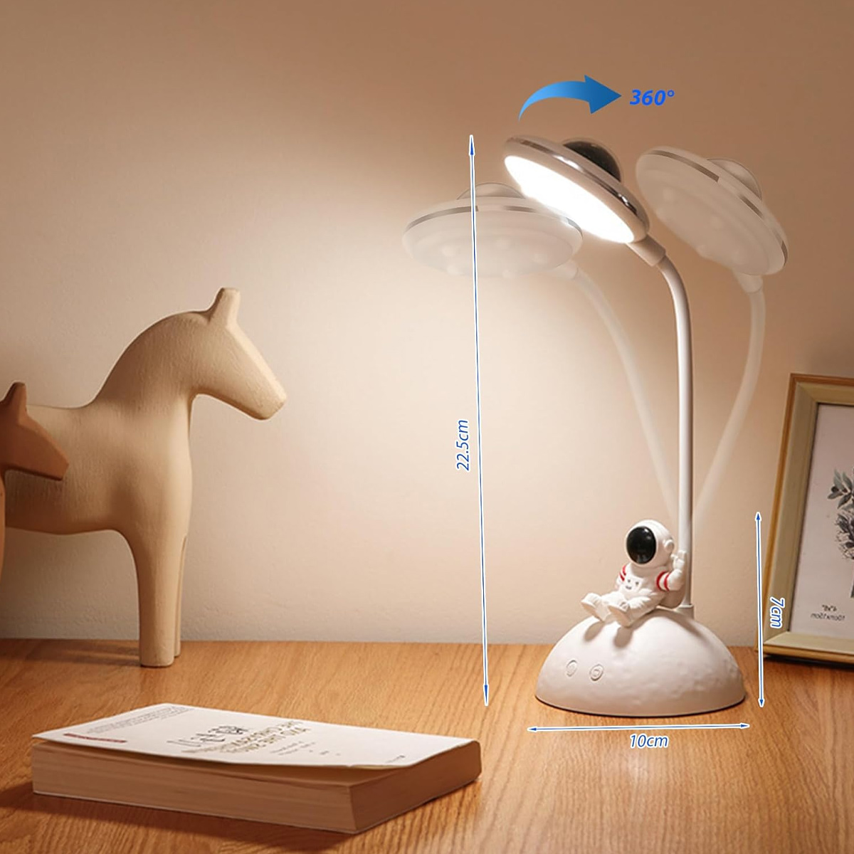 Eine weiße Astronautenlampe mit beweglichem Kopf auf einem Holztisch, daneben ein Buch und ein Holzpferd.