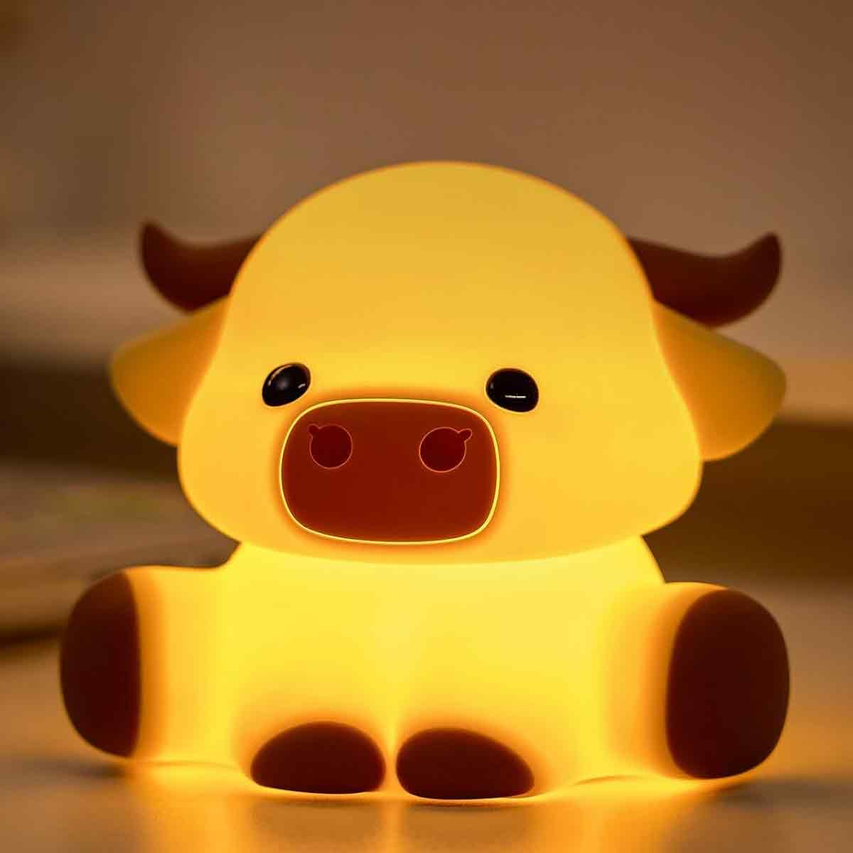 Eine niedliche, gelb leuchtende Nachtlampe in Form eines niedlichen Ochsen mit braunen Hörnern und Hufen sitzt auf einem Tisch.