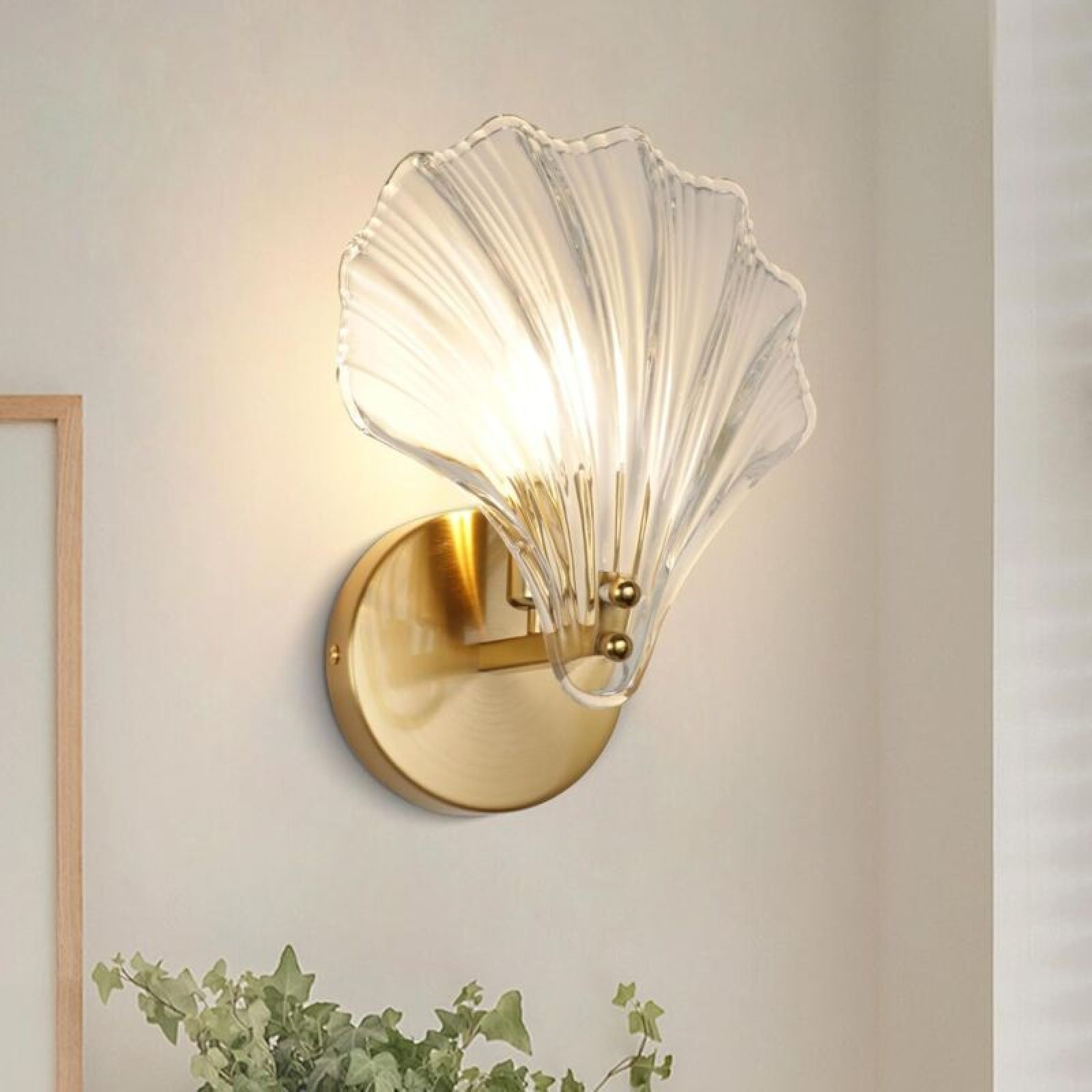 Eine goldene Wandleuchte mit einem gerippten, muschelförmigen Glasschirm leuchtet an einer hellen Wand.