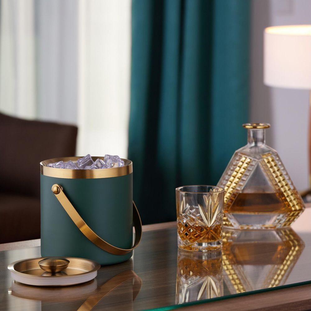 Ein grüner und goldener Eisbehälter, ein Whiskyglas und eine Karaffe stehen auf einem Glastisch.