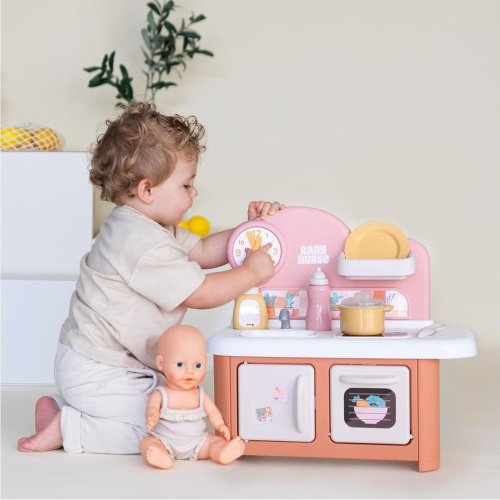 Ein kleiner Junge kniet vor einer rosa Spielküche und spielt mit einer Uhr, während eine Puppe daneben sitzt.
