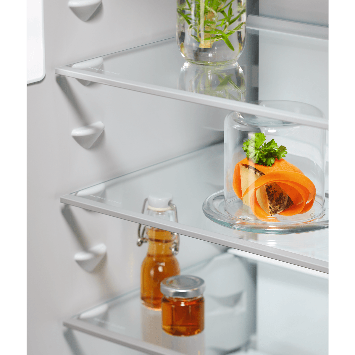 Estantes de vidrio en un refrigerador con hierbas frescas, un aperitivo bajo una campana y botellas.