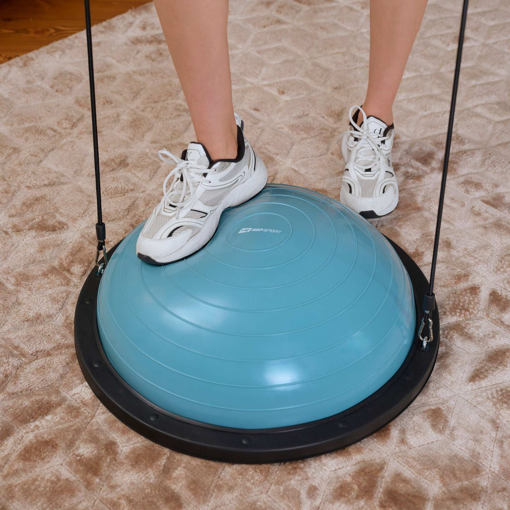 Person in weißen Turnschuhen steht auf blauem Balance-Trainer mit Widerstandsbändern auf Teppich.
