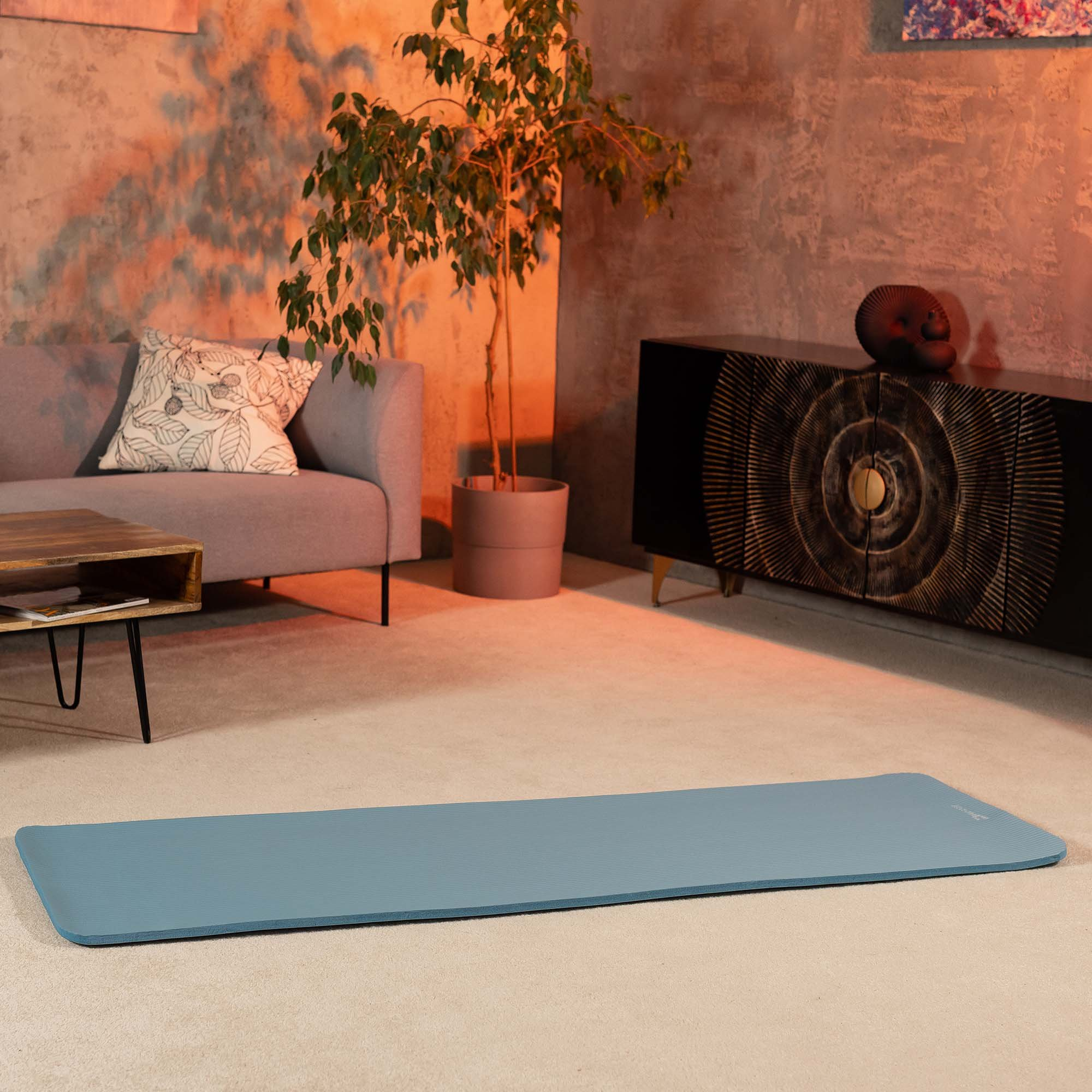 Eine blaue Yogamatte liegt auf dem Boden in einem warm beleuchteten Raum mit grauem Sofa, Pflanze und dunklem Sideboard.