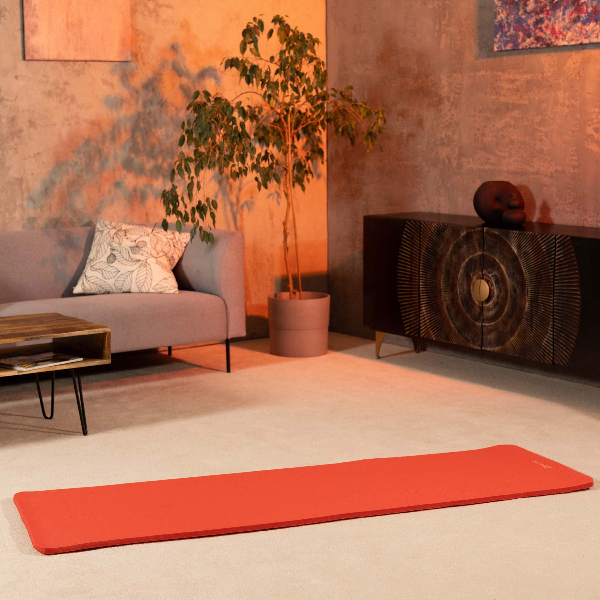 Eine rote Yogamatte liegt im Vordergrund eines Raumes mit grauer Couch, Holzmöbeln und einer Pflanze.