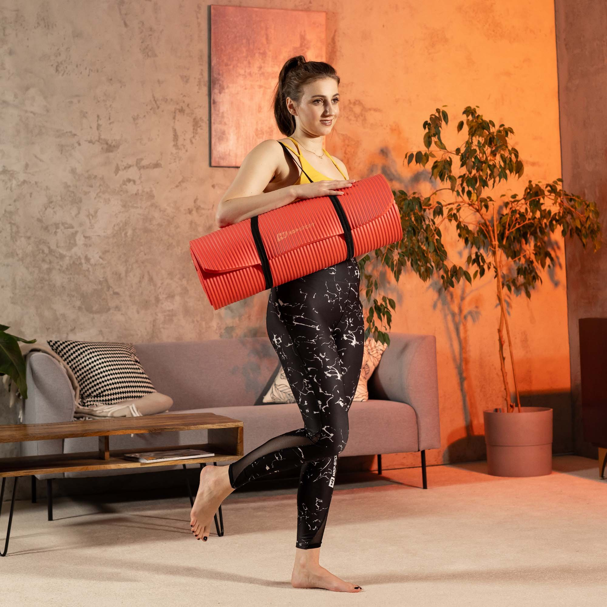 Eine Frau in Sportkleidung hält eine rote Fitnessmatte in einem gemütlichen Wohnzimmer.