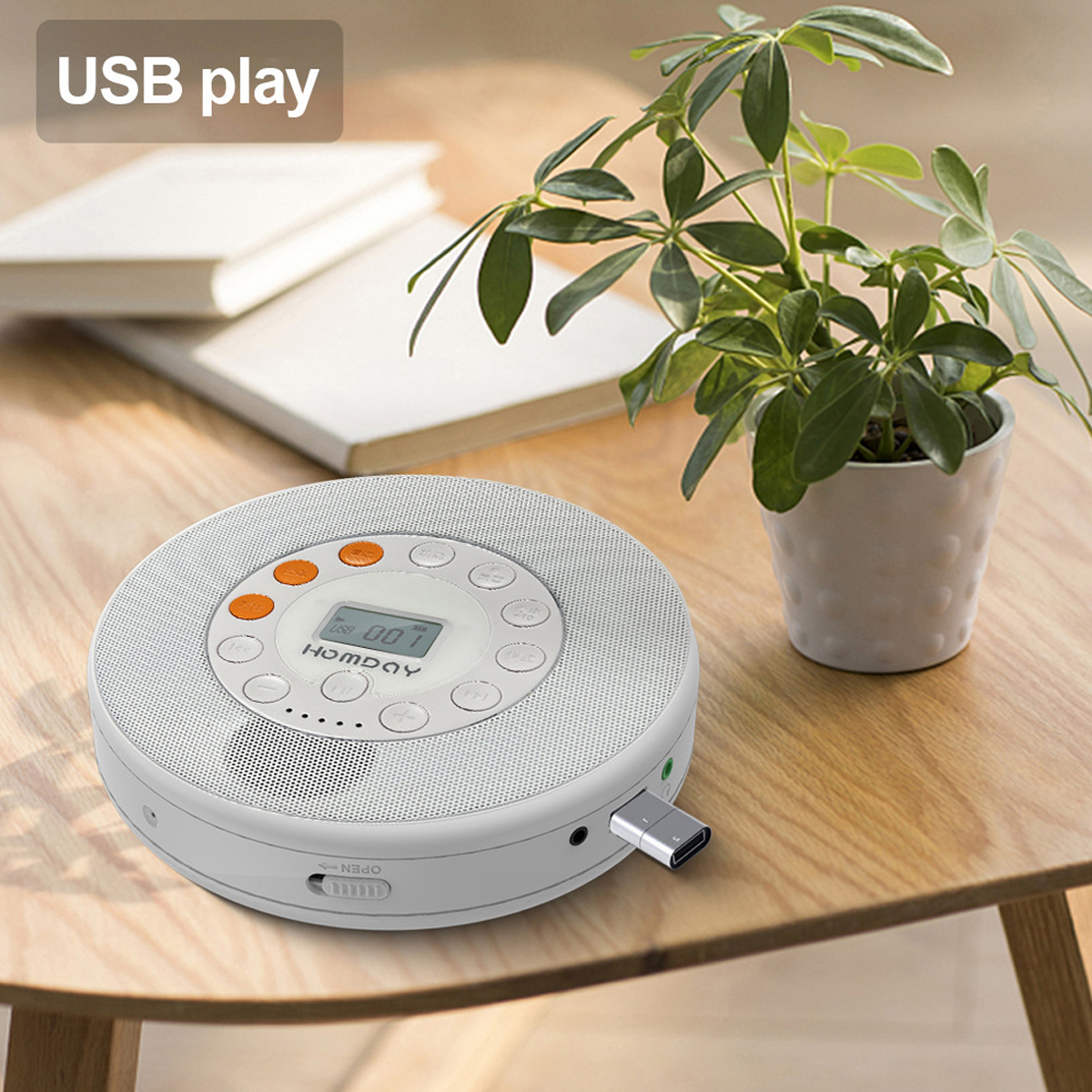 Ein runder weißer CD-Player auf einem Holztisch, daneben ein Buch und eine Topfpflanze.