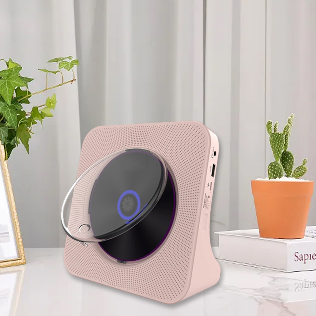 Ein rosafarbener CD-Player steht auf einem weißen Tisch zwischen einer Topfpflanze und einem kleinen Kaktus.