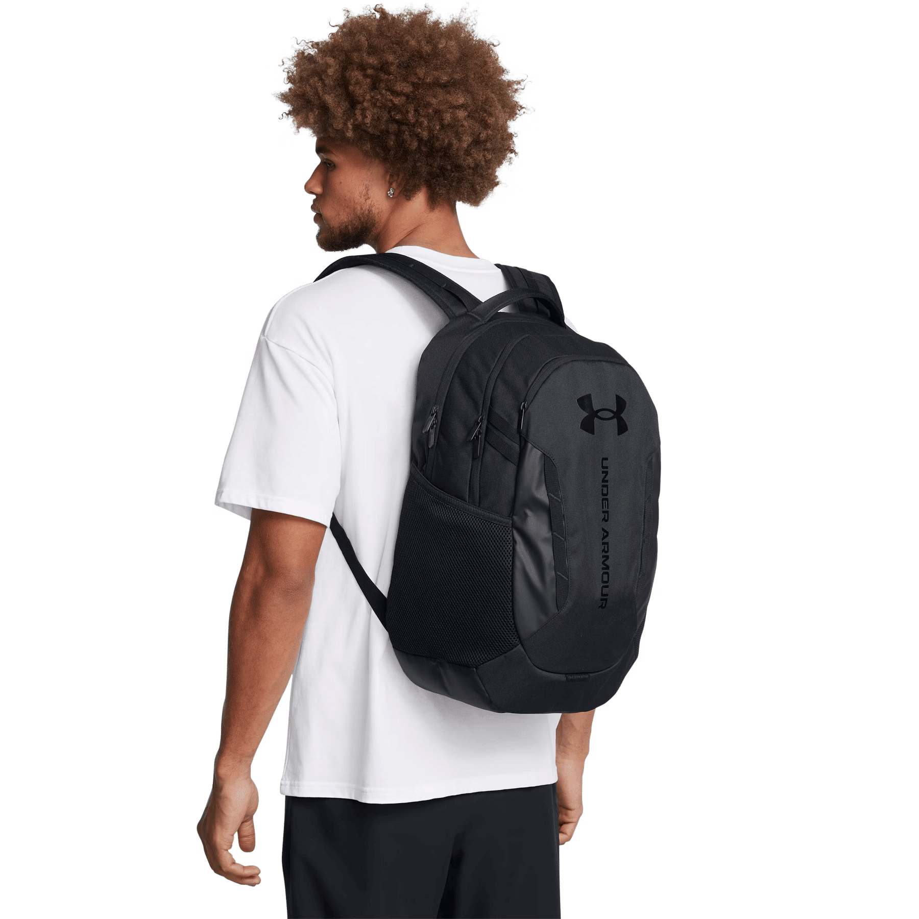 Ein lockiger Mann im weißen T-Shirt und schwarzen Shorts trägt einen schwarzen Under Armour Rucksack.