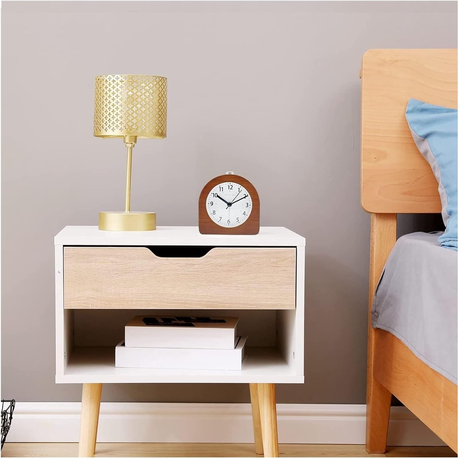 Ein weißer Nachttisch mit Holzakzent trägt eine goldene Lampe und Uhr, unten liegen zwei Bücher neben einem Holzbett.