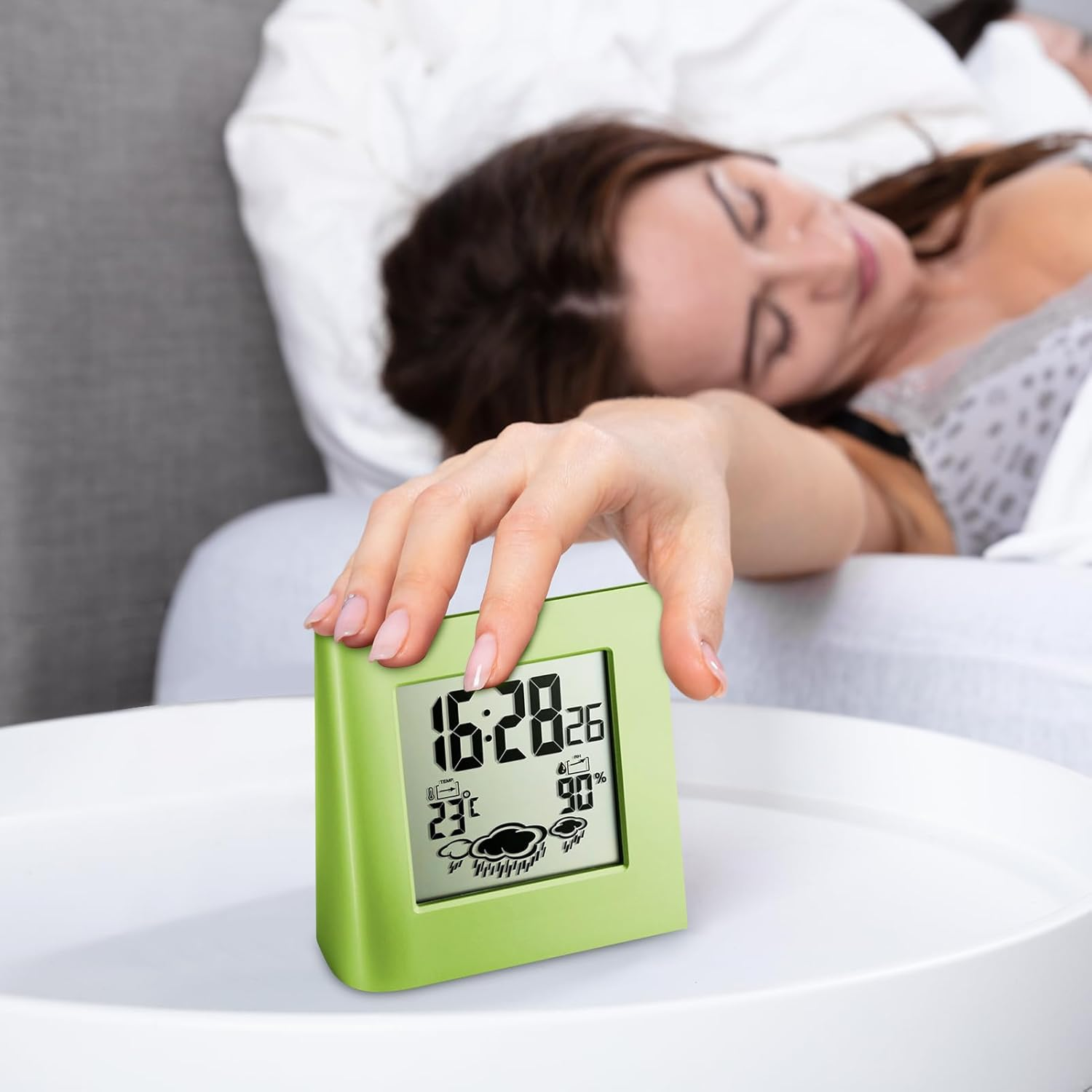 Eine Frau im Bett schaltet auf einem weißen Nachttisch einen grünen digitalen Wecker mit Uhrzeit und Wetter aus.