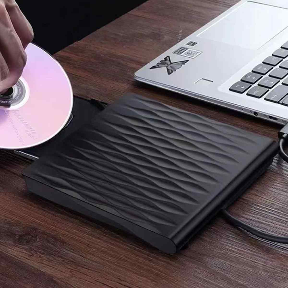 Eine Hand legt eine CD in ein externes schwarzes Laufwerk auf einem Holzschreibtisch neben einem Laptop.